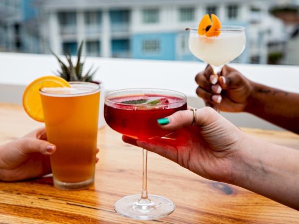 Three hands hold different drinks: a beer with an orange slice, a red cocktail, and a white drink with garnish, on a wooden table.