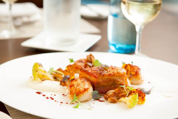 A gourmet dish featuring seared scallops, roasted vegetables, and sauce, elegantly plated with a glass of white wine in the background.