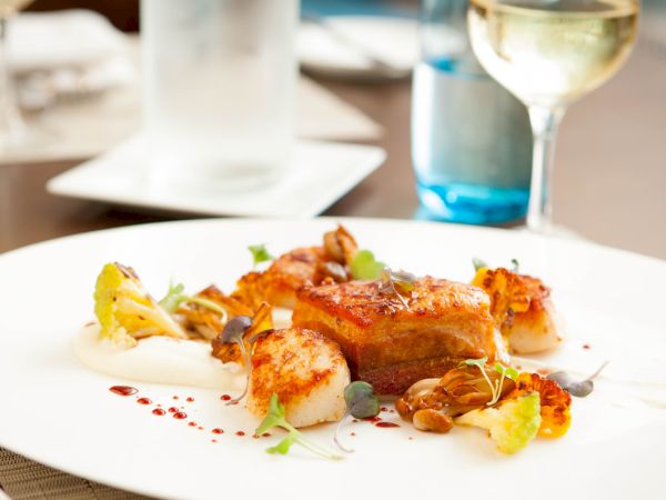 A gourmet dish featuring seared scallops, roasted vegetables, and sauce, elegantly plated with a glass of white wine in the background.