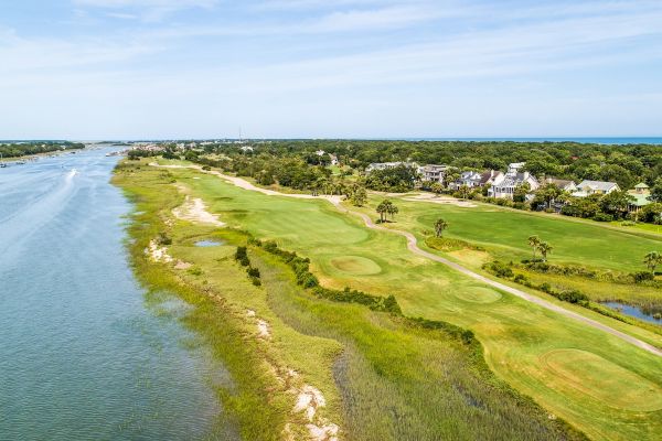 The image shows a scenic golf course beside a river, with houses and greenery in the background under a clear blue sky.