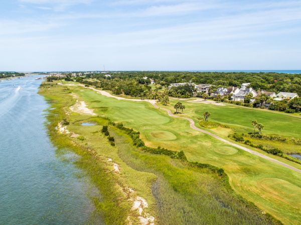 The image shows a scenic golf course beside a river, with houses and greenery in the background under a clear blue sky.