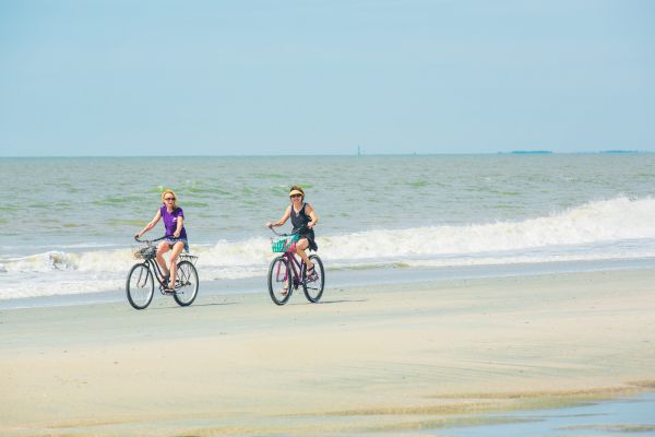 Two people are riding bicycles on a sandy beach near the ocean, with waves in the background and a clear sky above the scene.