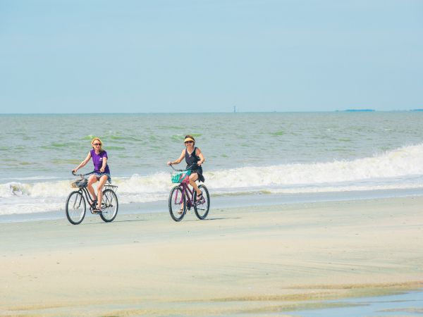 Two people are riding bicycles on a sandy beach near the ocean, with waves in the background and a clear sky above the scene.