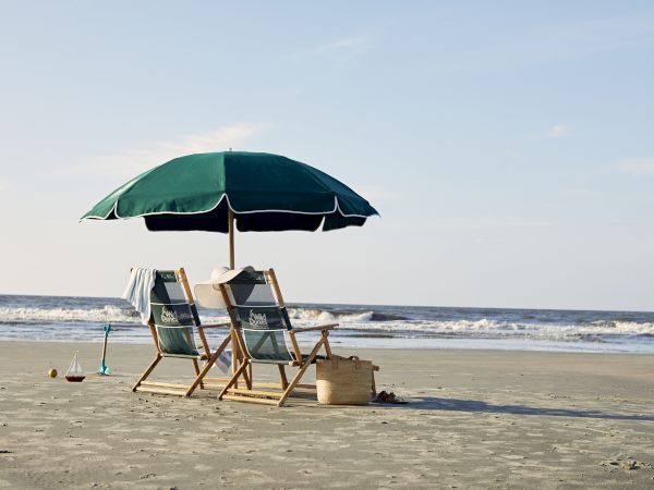 Two beach chairs under a green umbrella on sandy shore, with waves in the background and beach items nearby.