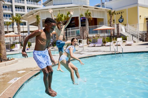 Three kids are joyfully jumping into a swimming pool, with a hotel in the background and palm trees.