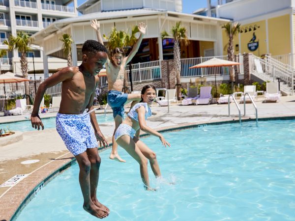 Three kids are joyfully jumping into a swimming pool, with a hotel in the background and palm trees.