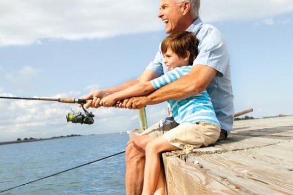 A man and a child are fishing together, sitting on a wooden pier overlooking the water, enjoying a sunny day.
