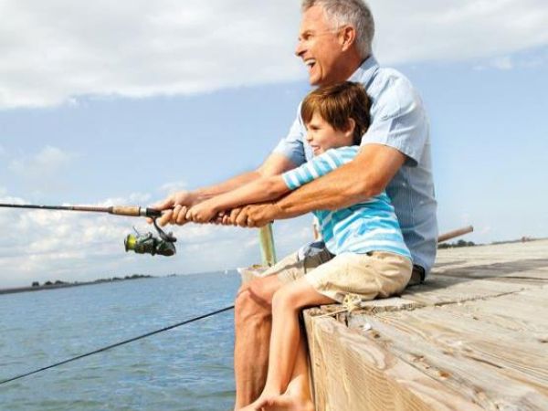 A man and a child are fishing together, sitting on a wooden pier overlooking the water, enjoying a sunny day.
