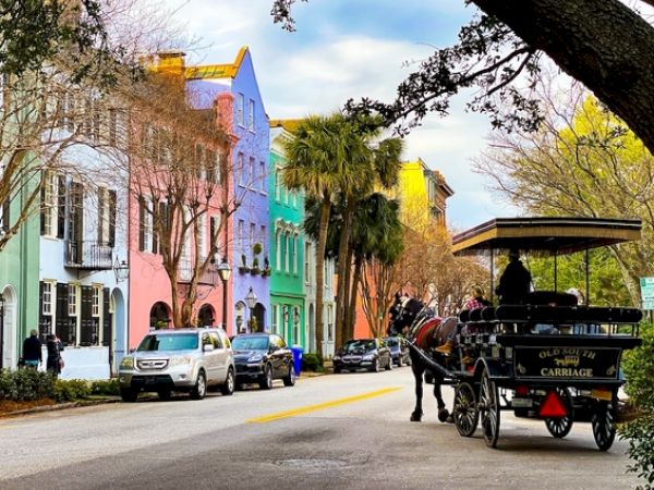 A colorful street with pastel buildings, parked cars, and a horse-drawn carriage under tree-lined surroundings.
