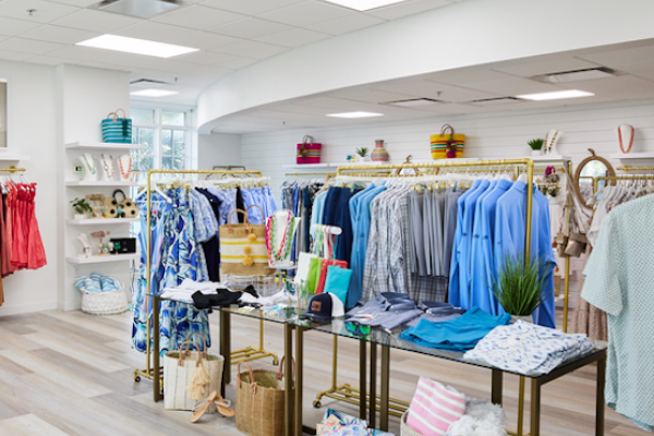The image shows a clothing store interior with racks of colorful clothes and accessories, neatly displayed on wooden floors with bright lighting.
