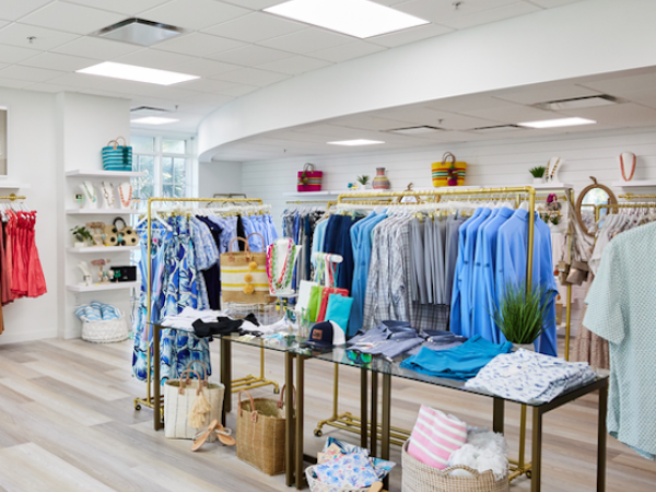 The image shows a clothing store interior with racks of colorful clothes and accessories, neatly displayed on wooden floors with bright lighting.