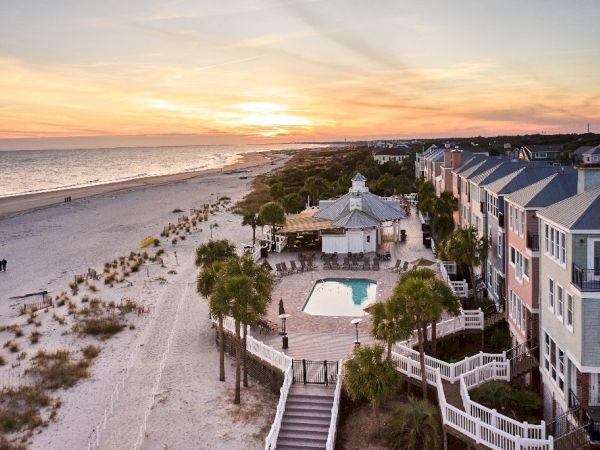 A serene beach scene at sunset, with a pool, palm trees, and colorful buildings lined along the shore.
