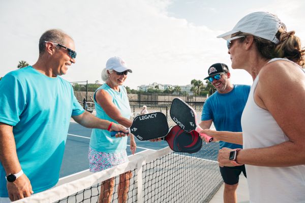 Four people are on a pickleball court, tapping paddles over the net in a friendly gesture, wearing casual sports attire.