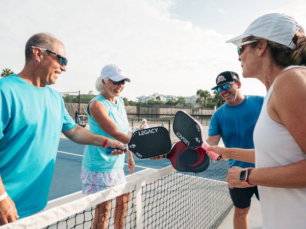 Four people are on a pickleball court, tapping paddles over the net in a friendly gesture, wearing casual sports attire.
