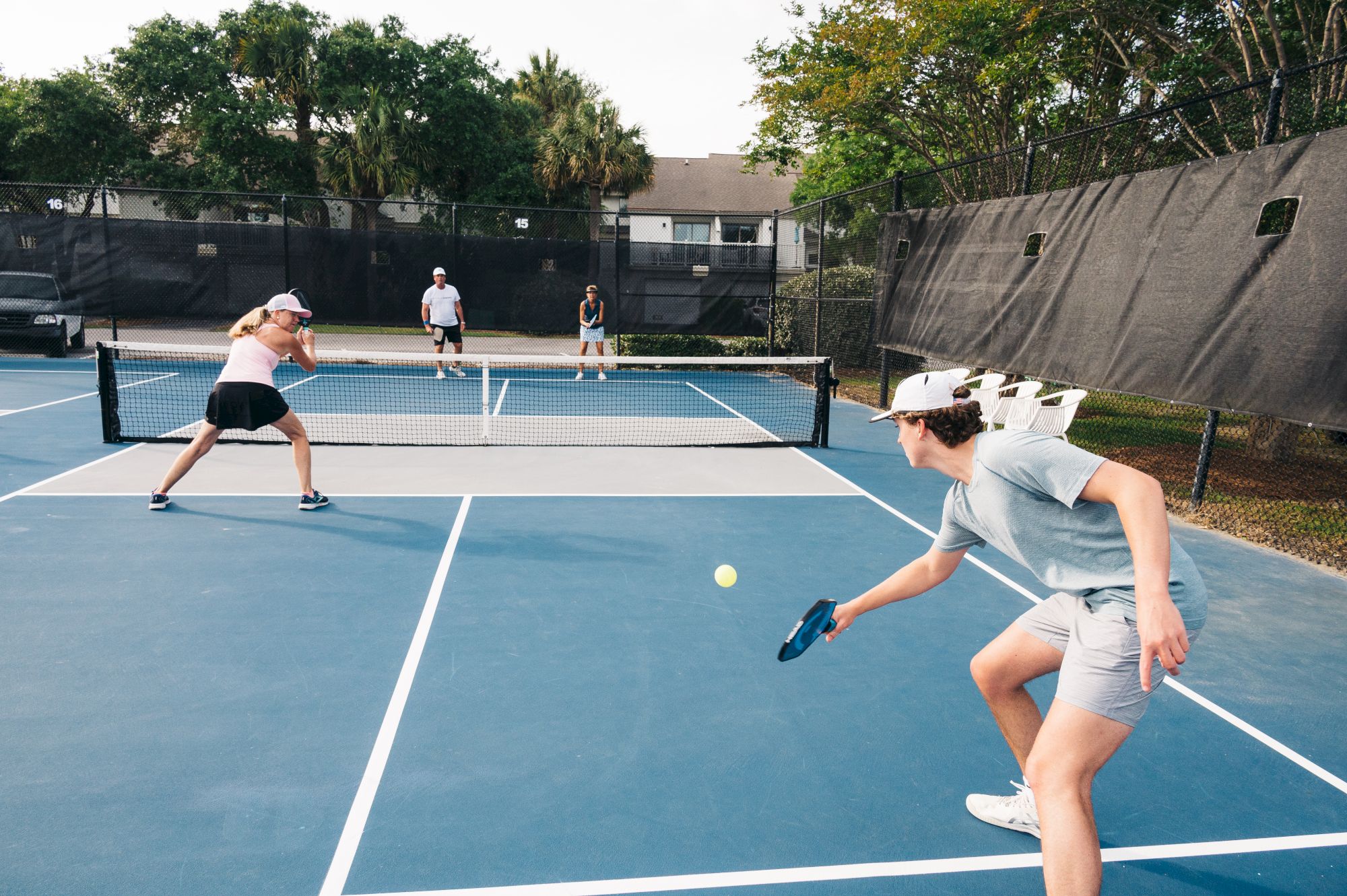 People are playing pickleball on an outdoor court, with one player hitting the ball while others observe in the background.