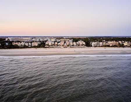 A coastal landscape showing the ocean, a sandy beach, and a row of beachfront buildings under a pastel sky at sunset or sunrise.