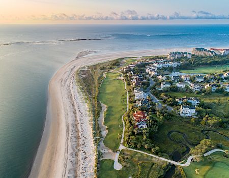A coastal aerial view features a sandy beach, lush greenery, residential buildings, and a golf course by the ocean under a clear sky.
