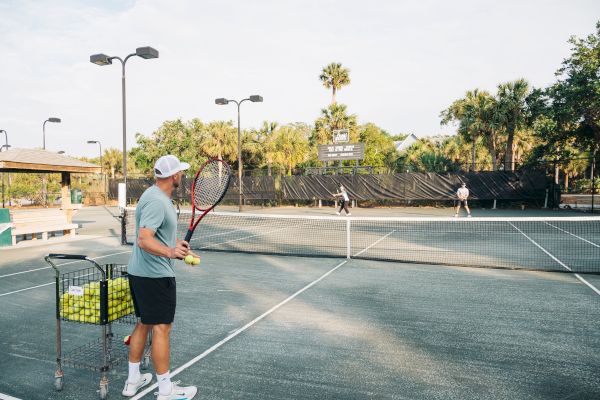 A person on a tennis court holding a racket and tennis balls, with a cart full of balls nearby, observing two players.