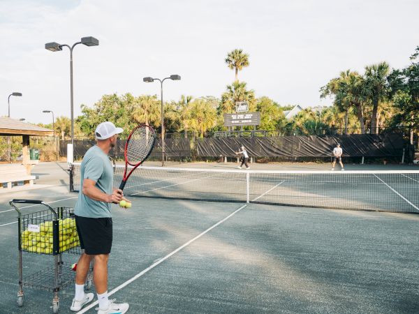 A person on a tennis court holding a racket and tennis balls, with a cart full of balls nearby, observing two players.