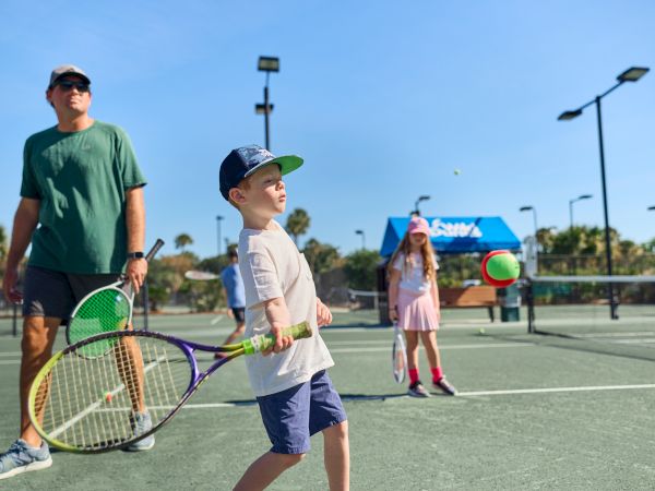 A father watches as a boy in a cap serves a tennis ball on an outdoor court, with teammates in the background under a sunny, blue sky.