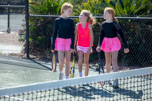 Three young girls in tennis outfits walk on a court, holding rackets, and chatting. They're wearing black tops and pink skirts.