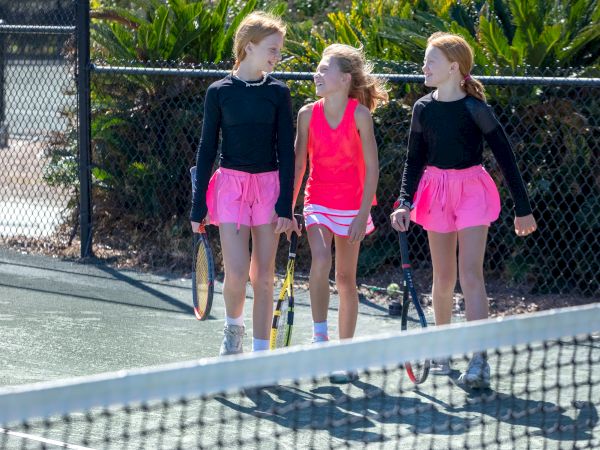 Three young girls in tennis outfits walk on a court, holding rackets, and chatting. They're wearing black tops and pink skirts.