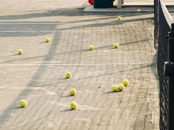 Tennis balls scattered on a court near a fence, some shadows visible on the ground.