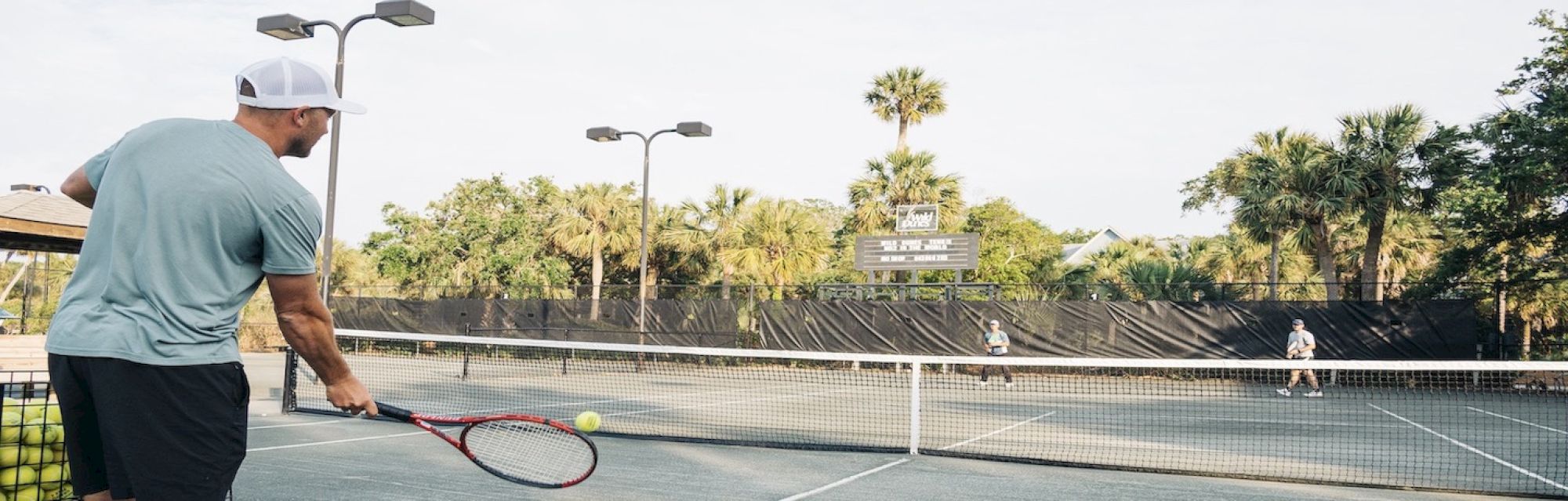 A person serves a tennis ball on an outdoor court amidst palm trees, with two players visible on the opposite side of the net.