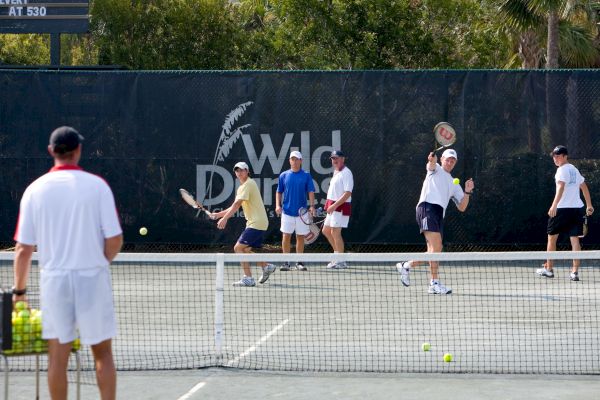 A group of people are playing tennis on an outdoor court, with a coach watching. There's a sign in the background reading "Wild Dunes."