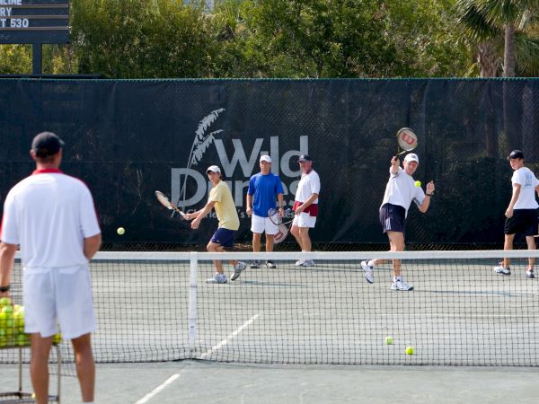 A group of people are playing tennis on an outdoor court, with a coach watching. There's a sign in the background reading "Wild Dunes."