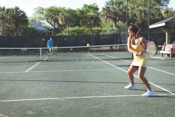 Two people playing tennis on an outdoor court, surrounded by trees and a fence, with tennis balls scattered on the ground.