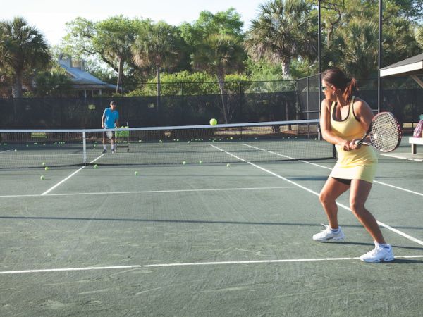Two people playing tennis on an outdoor court, surrounded by trees and a fence, with tennis balls scattered on the ground.