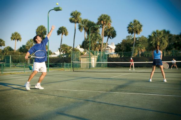 People playing tennis on an outdoor court surrounded by palm trees under a clear blue sky.