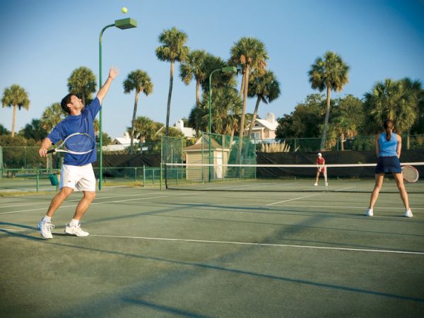 People playing tennis on an outdoor court surrounded by palm trees under a clear blue sky.