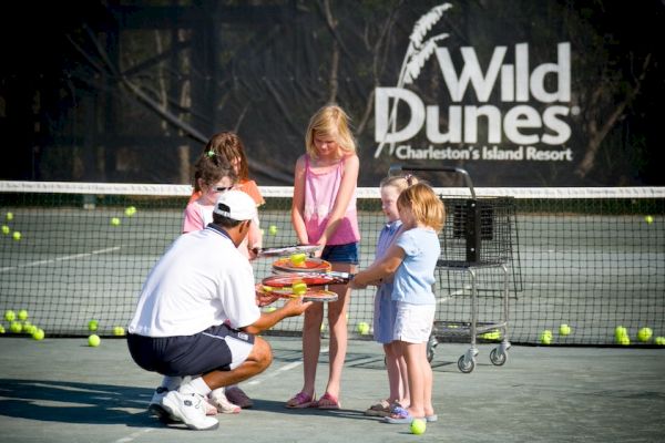 A tennis coach is handing rackets to a group of children on a court, with the Wild Dunes resort sign in the background.
