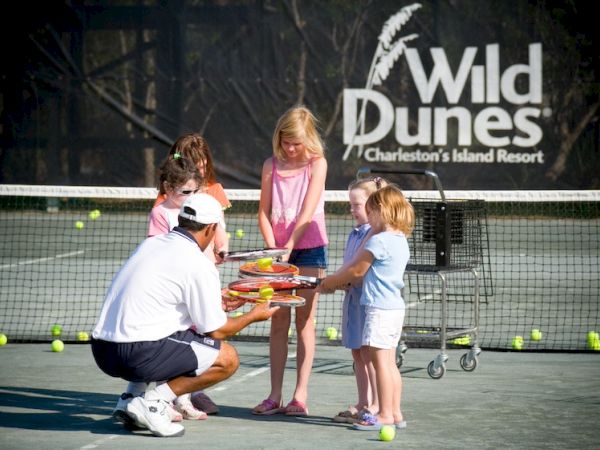 A tennis coach is handing rackets to a group of children on a court, with the Wild Dunes resort sign in the background.