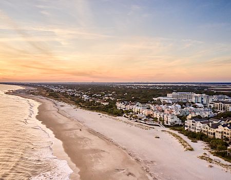 A scenic coastal view with a sandy beach, calm ocean waves, and buildings along the shore under a colorful sunset sky.