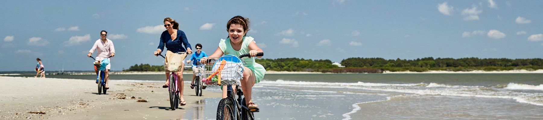 People are riding bicycles along a sandy beach with waves in the background, under a clear blue sky.