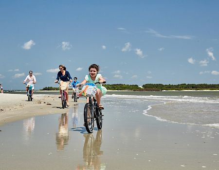 People are riding bicycles along a sandy beach with waves in the background, under a clear blue sky.