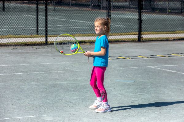 A young girl stands on a tennis court holding a tennis racket, wearing a blue shirt and pink leggings.