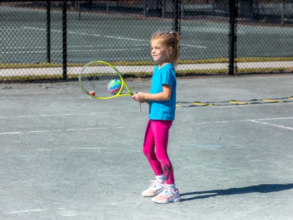 A young girl stands on a tennis court holding a tennis racket, wearing a blue shirt and pink leggings.