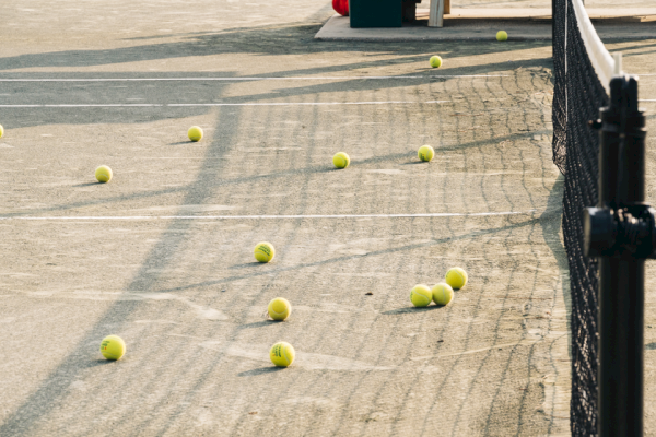 A tennis court with several tennis balls scattered around, situated next to a net under sunlight, creates a playful scene.