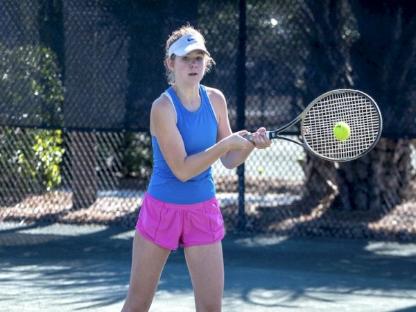 A person is playing tennis on an outdoor court, wearing a blue tank top and pink shorts, preparing to hit the ball with a backhand swing.