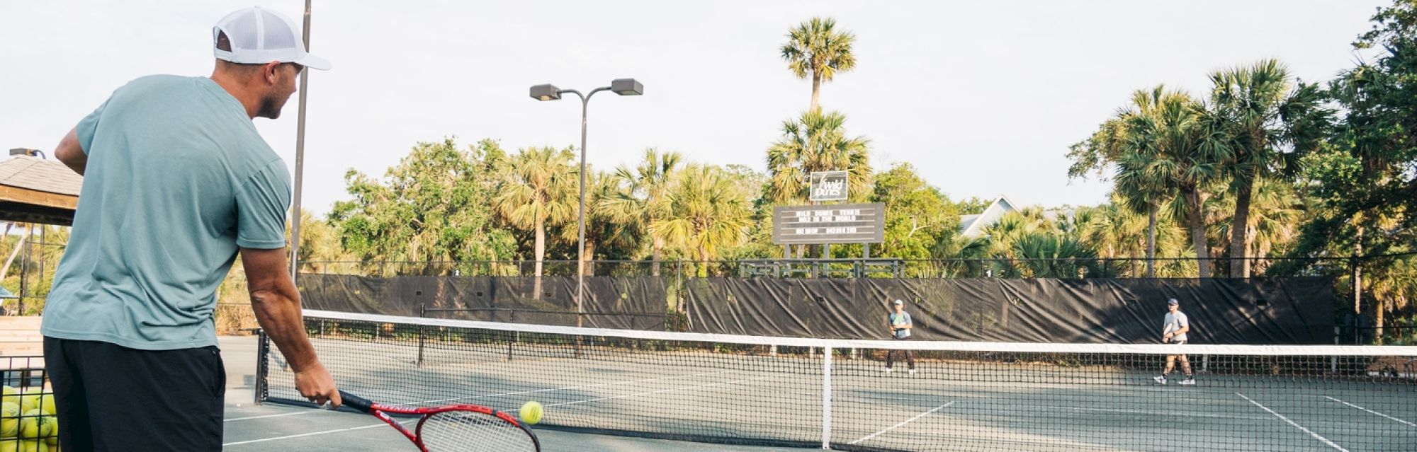 A person plays tennis on an outdoor court surrounded by palm trees, facing an opponent across the net.