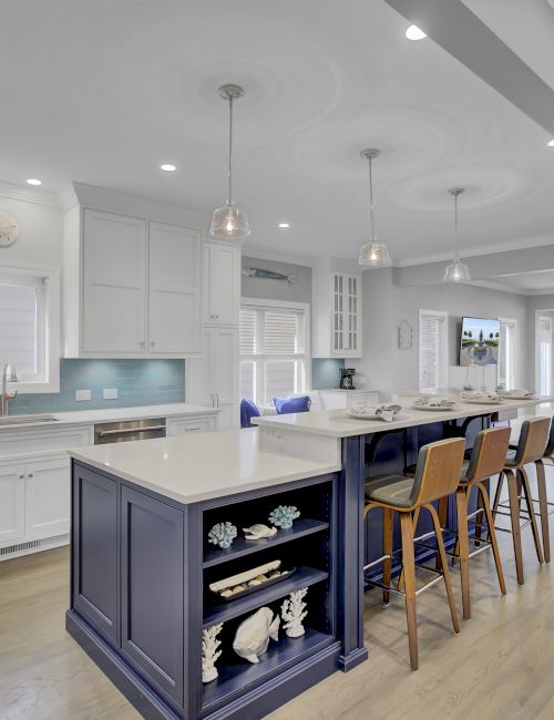 A modern kitchen with white cabinets, a blue island, bar stools, and pendant lights, adjacent to a spacious open living area.