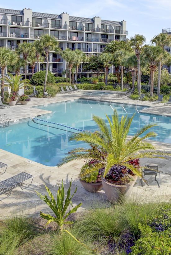 The image shows an outdoor swimming pool surrounded by lounge chairs, plants, and a multi-story residential building in the background.