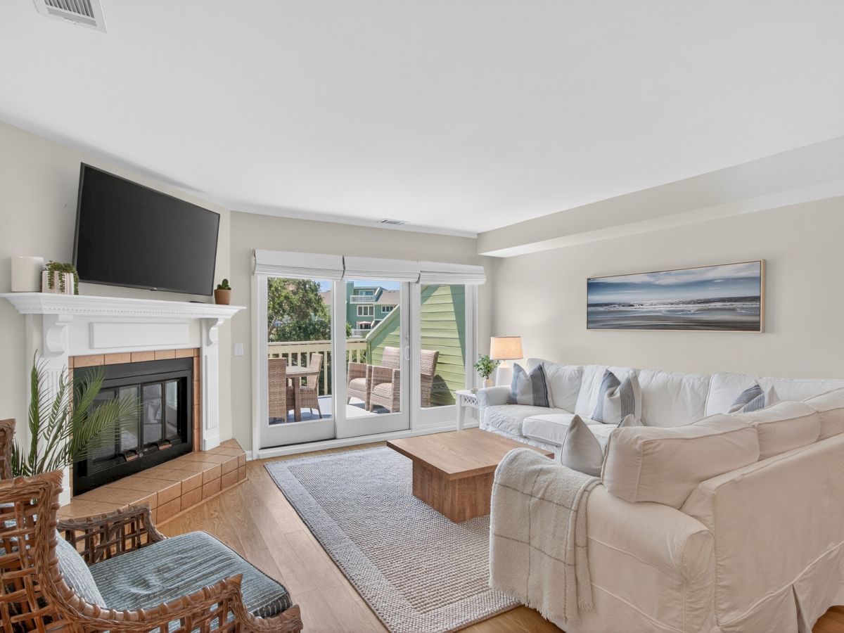 A cozy living room features a white sectional sofa, fireplace, wall-mounted TV, and a view of an outdoor deck through glass doors.