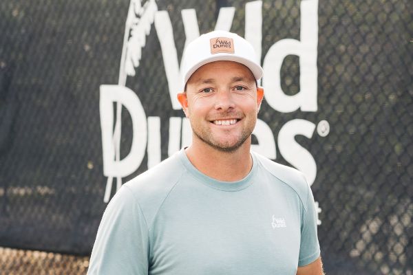 A person wearing a cap and t-shirt stands in front of a "Wild Dunes" sign, smiling at the camera.