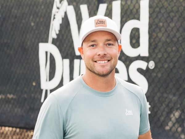 A person wearing a cap and t-shirt stands in front of a "Wild Dunes" sign, smiling at the camera.
