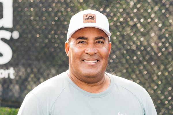 A person wearing a white "Wild Dunes" cap and light gray shirt smiles against a blurred outdoor background.